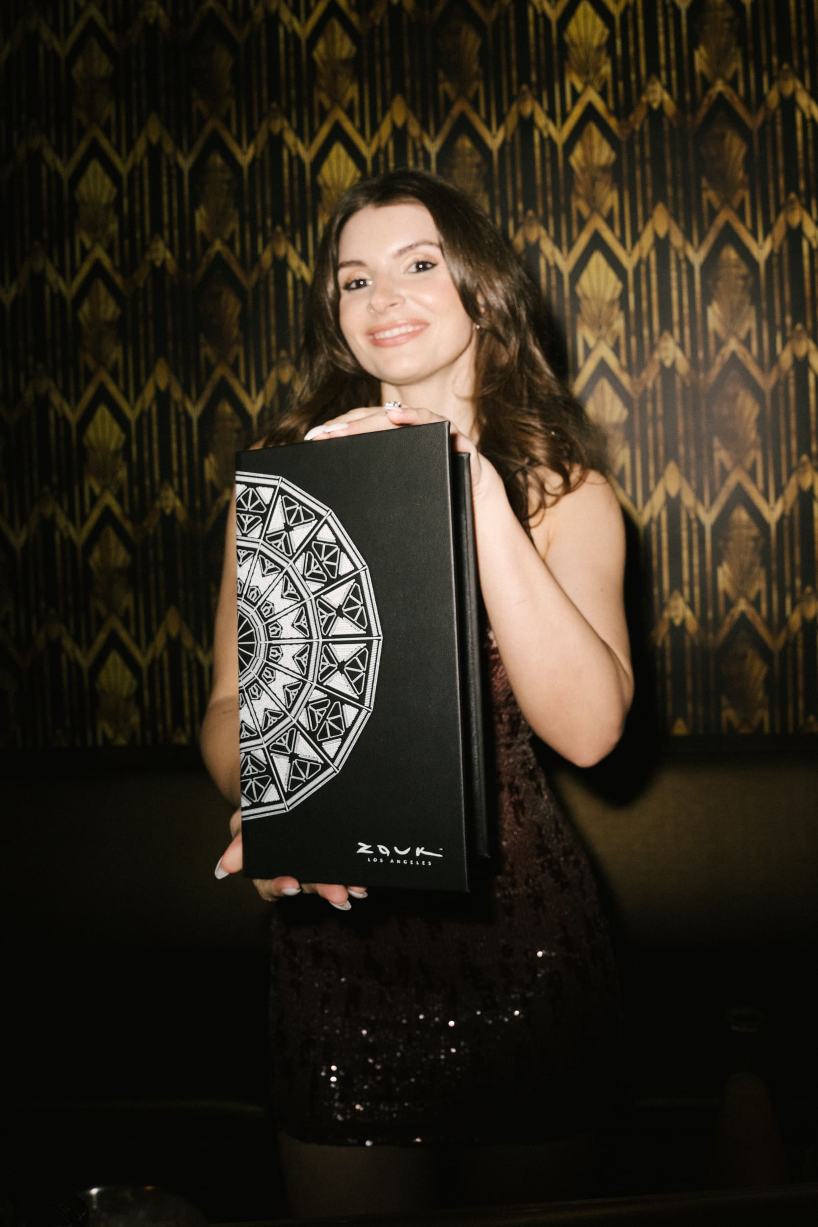A woman in a shimmering dress is smiling and holding a box with a detailed design, showcasing the "Zouk" logo. The background features an elegant patterned wall, adding a vibrant touch to the scene.