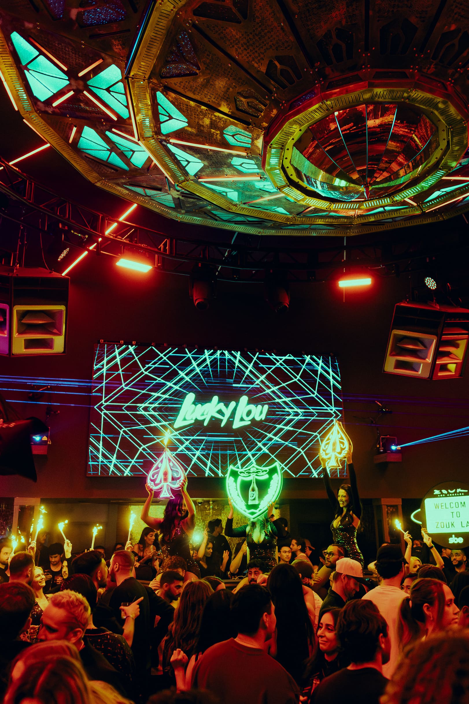 Crowd enjoying a nightclub performance under a colorful LED ceiling with dancers holding neon signs and "Lucky Lou" displayed on screen.