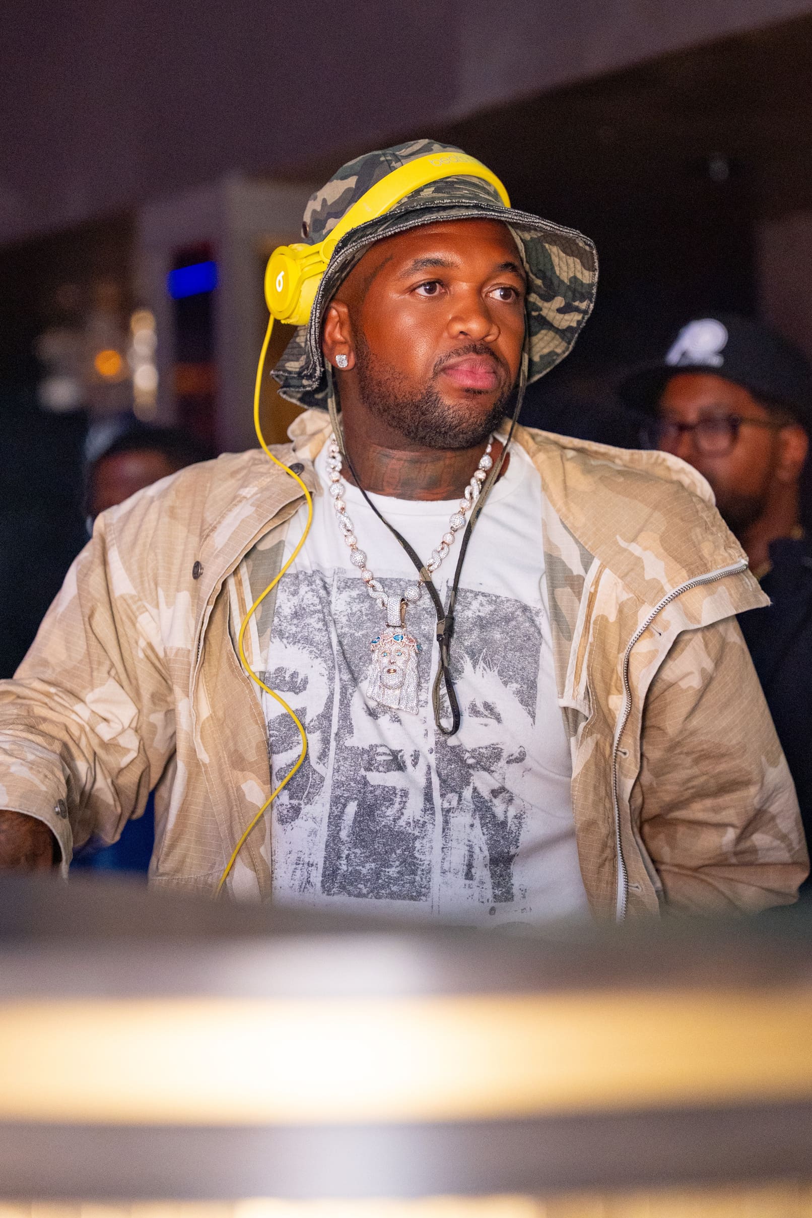 Man wearing camo jacket and bucket hat with yellow headphones around his neck at a nightclub.