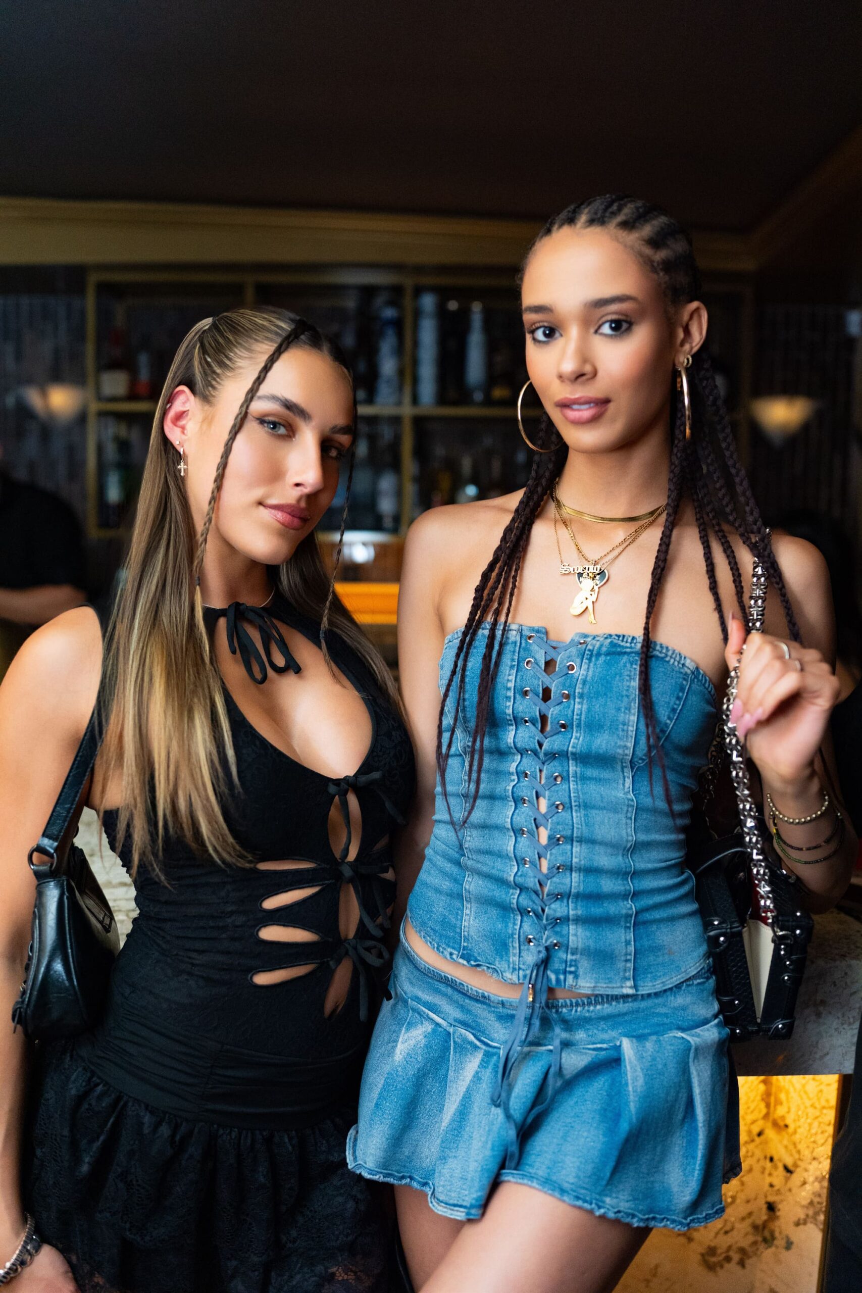Two women posing together inside a nightclub, one wearing a black cutout dress and the other in a denim corset top and skirt.
