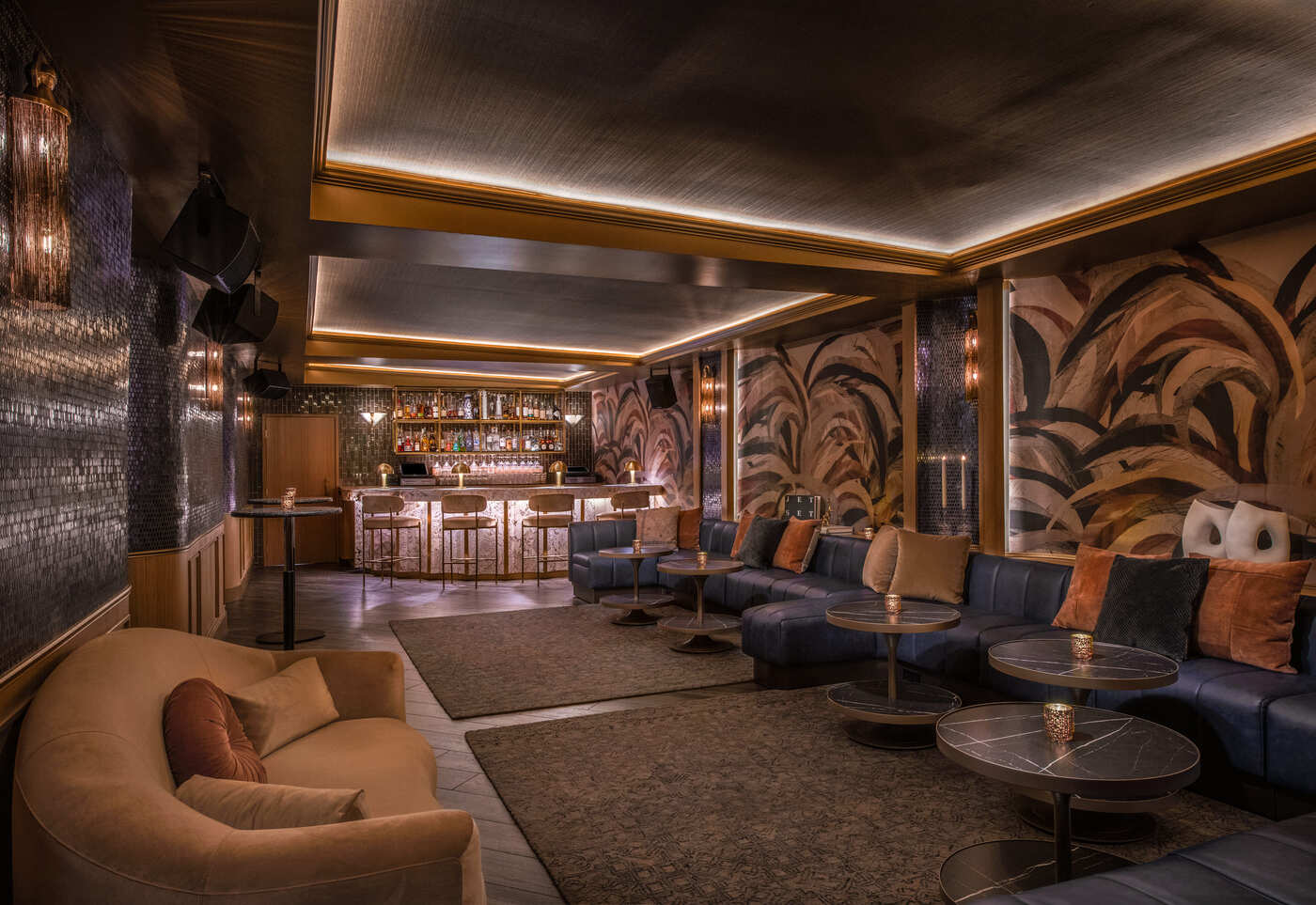 Upscale lounge interior with curved velvet seating, textured walls, ambient lighting, and a marble-topped bar in the background.
