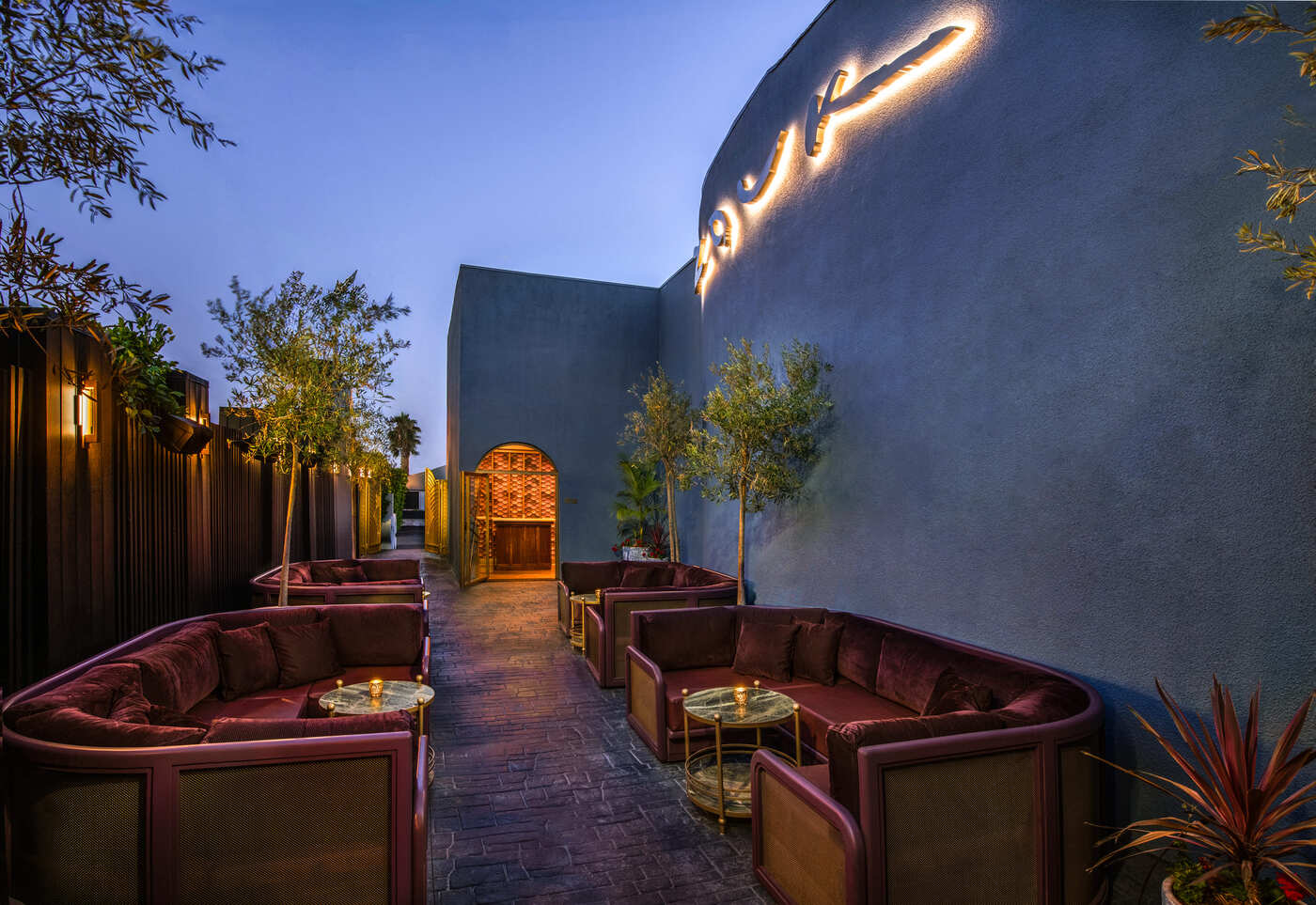 Outdoor lounge area with plush maroon seating, small round tables, and decorative trees along a stone path leading to a wooden door.