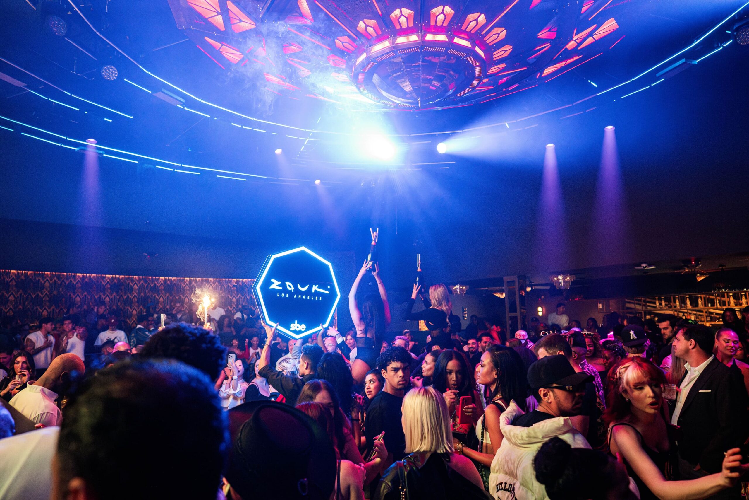 Nightclub crowd dancing under vibrant lights with performers holding a Zouk Los Angeles sign and champagne sparklers.