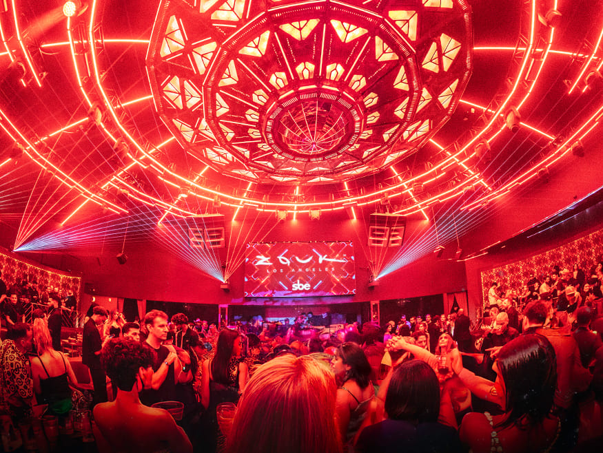 Crowded nightclub with red lighting, ceiling lasers, and a central dome fixture above the dance floor.