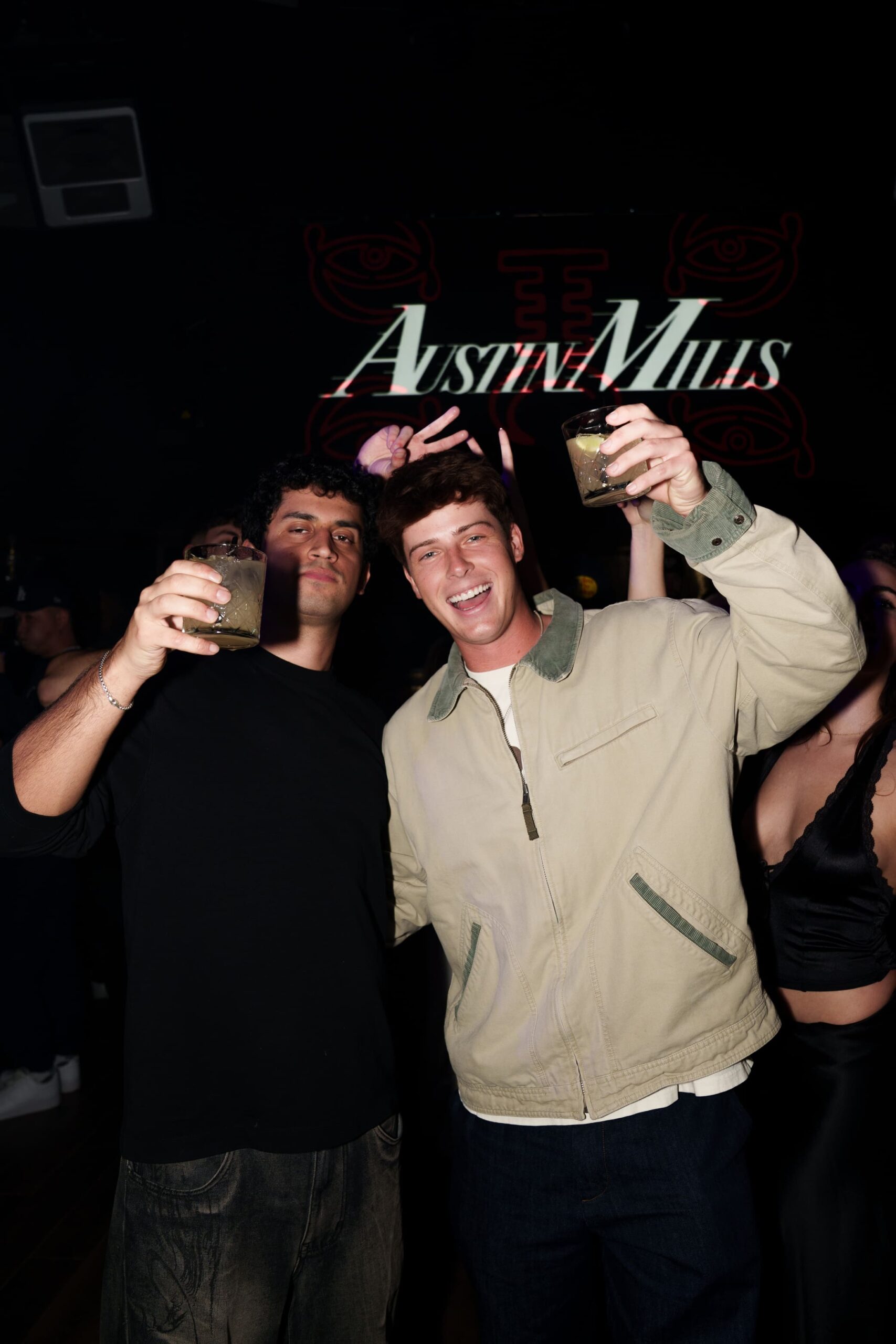 Two men smiling and holding drinks at a nightclub with “Austin Mills” displayed on the screen behind them.