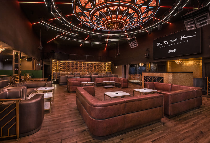 A stylish nightclub lounge interior featuring U-shaped brown leather sofas with small marble tables, warm wooden flooring, a decorative illuminated ceiling fixture, and a “ZOUK Los Angeles” sign on the back wall.