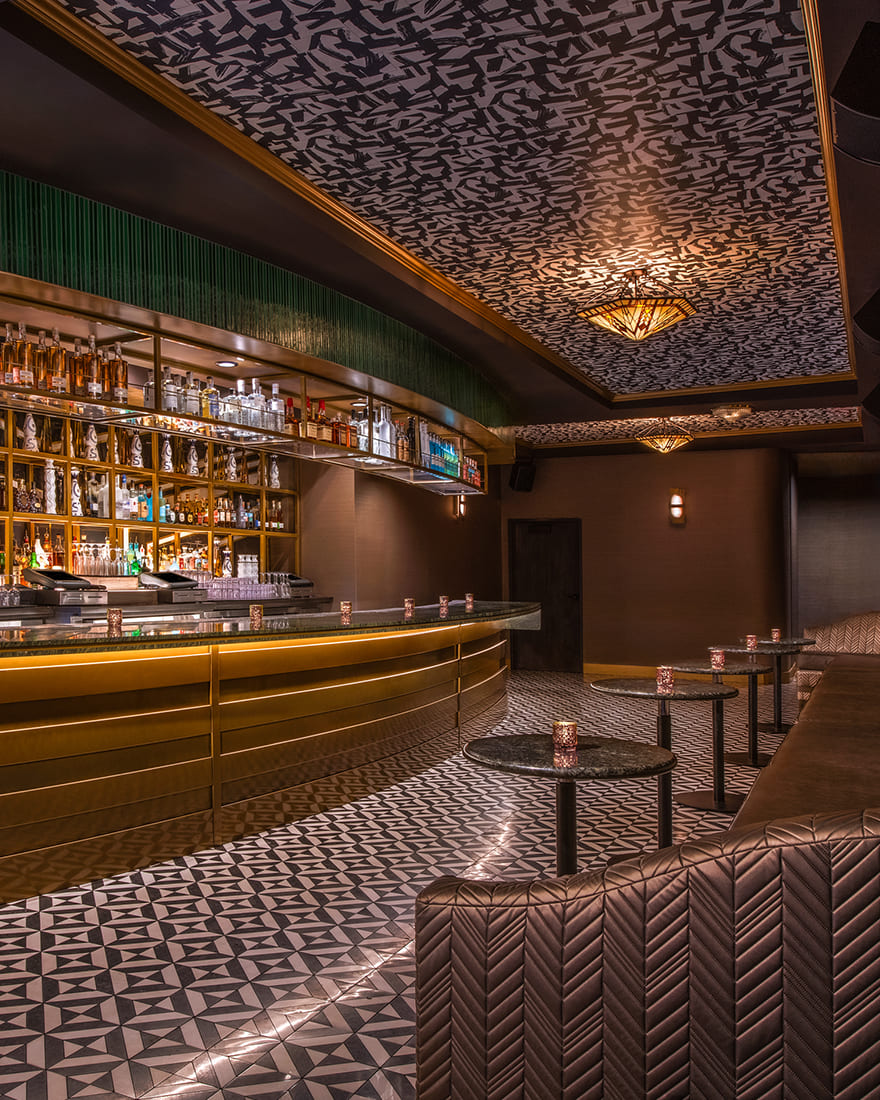Elegant bar interior with a curved brass counter, patterned tile flooring, geometric ceiling design, and leather booth seating with small round tables and votive candles.