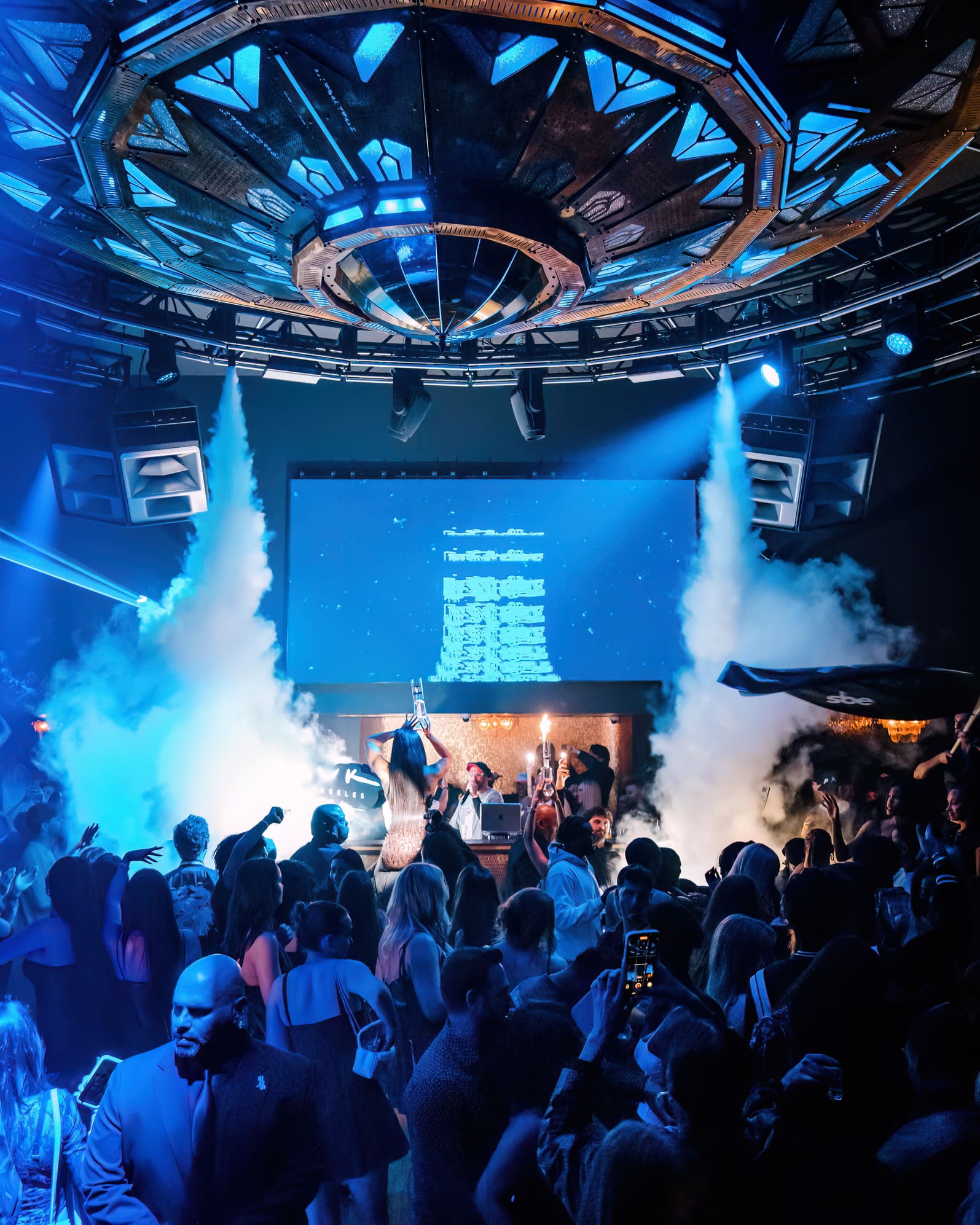 Crowd enjoying a nightclub party with blue lighting, smoke effects, and illuminated ceiling installation above the DJ booth.