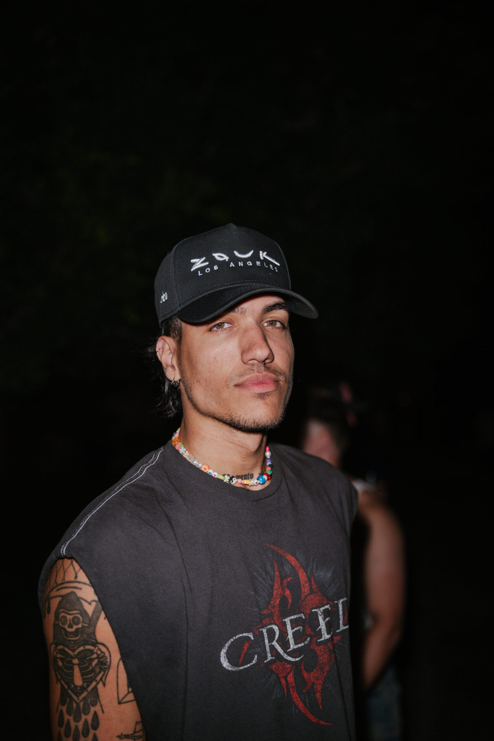 Man wearing a Zouk Los Angeles hat and Creed t-shirt poses at night with a dark background.