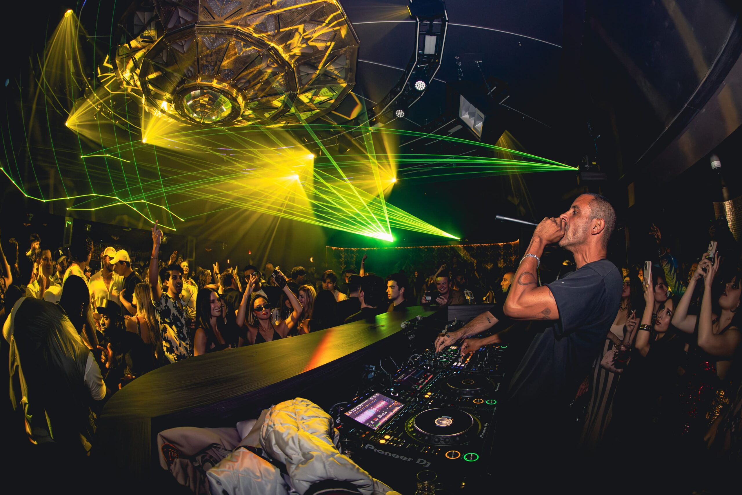 DJ performing in front of a lively crowd with green and yellow laser lights under a large decorative ceiling fixture.