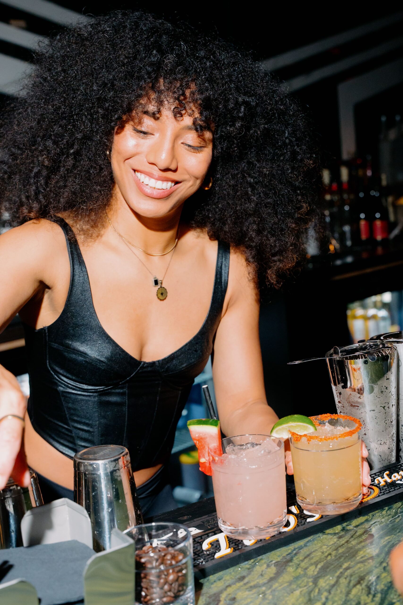 Smiling bartender in a black top serving two colorful cocktails at a dimly lit bar.