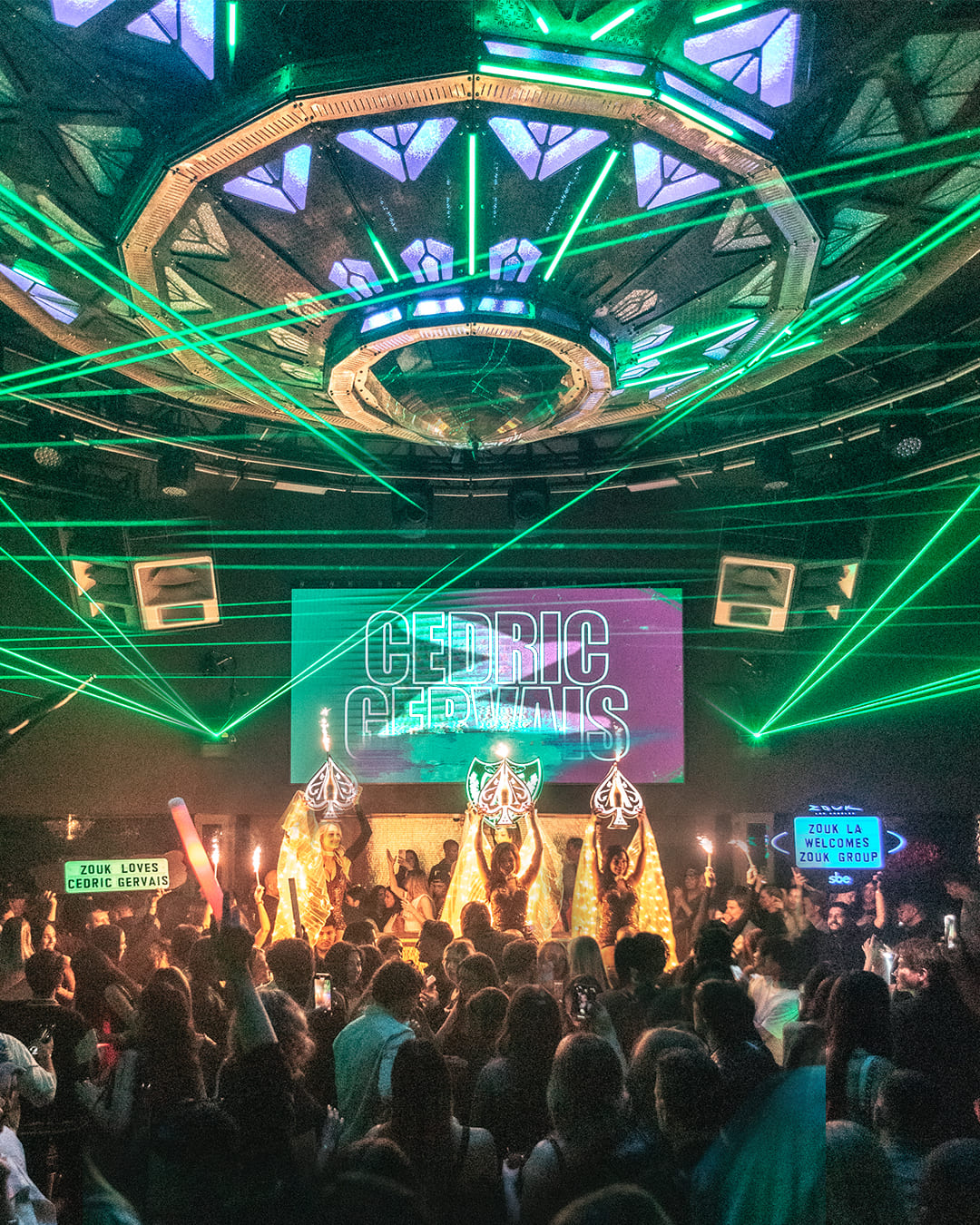 Crowded nightclub with green laser lights, performers holding light displays, and “Cedric Gervais” displayed on a large screen.