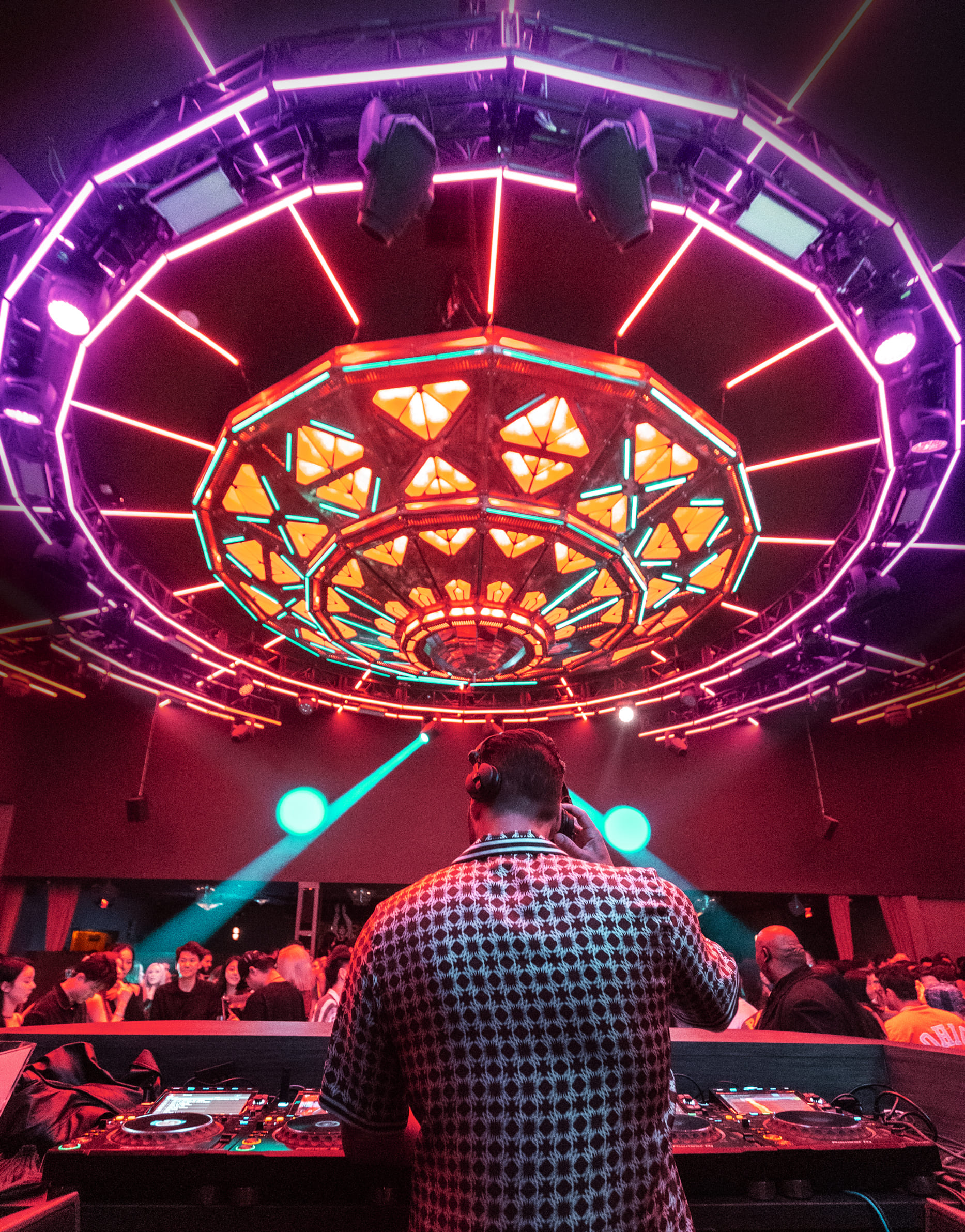 DJ performing in a nightclub with a colorful, illuminated ceiling and a crowd dancing below.