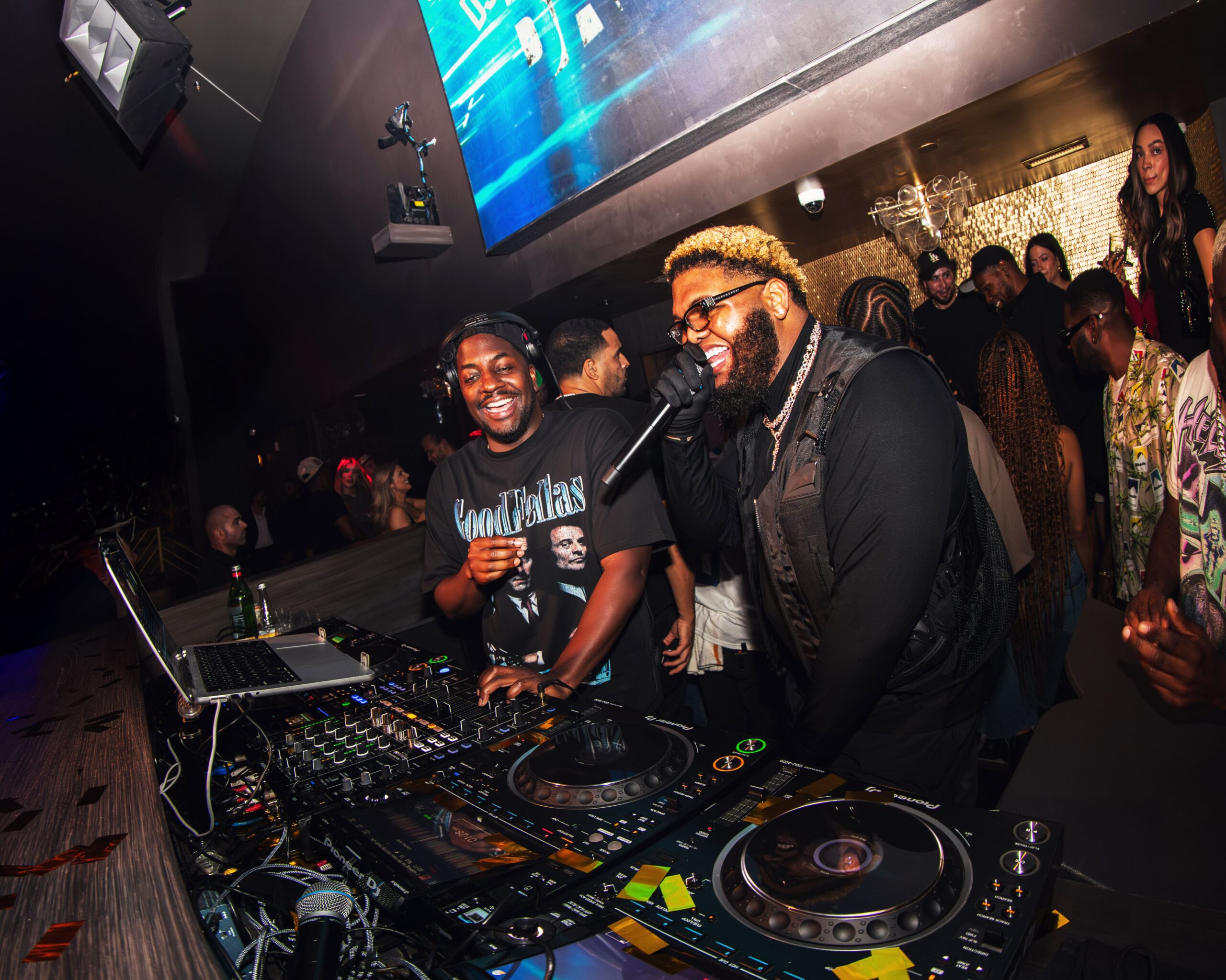 Two DJs performing at a nightclub booth, one speaking into a microphone while the other smiles.