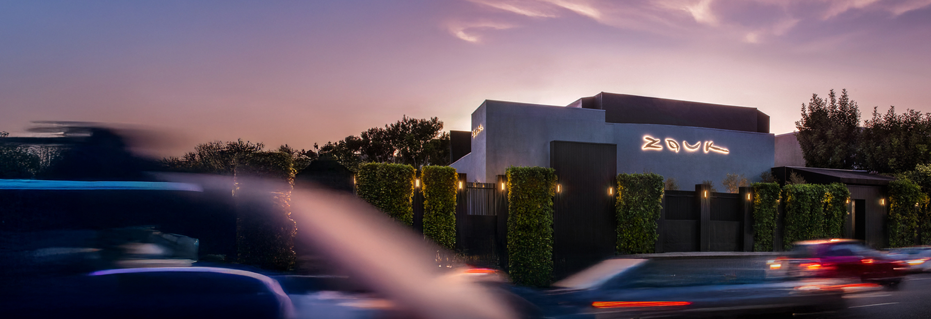 Exterior view of Zouk Los Angeles at dusk, with cars passing by and illuminated signage on a modern building facade.